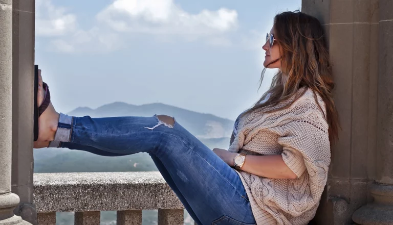 uma mulher, de calças de ganga e blusa branca, sentada junto a uma varanda, usando chinelos e óculos de sol, demonstrando que está a viajar, aproveitando o ano sabático