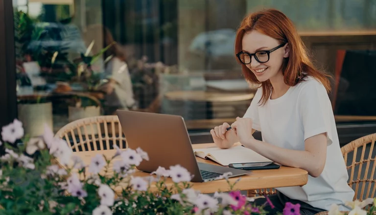 Uma mulher de cabelo ruivo, com óculos, sentada numa mesa exterior, em frente a um computador, a aprender através de cursos à distância
