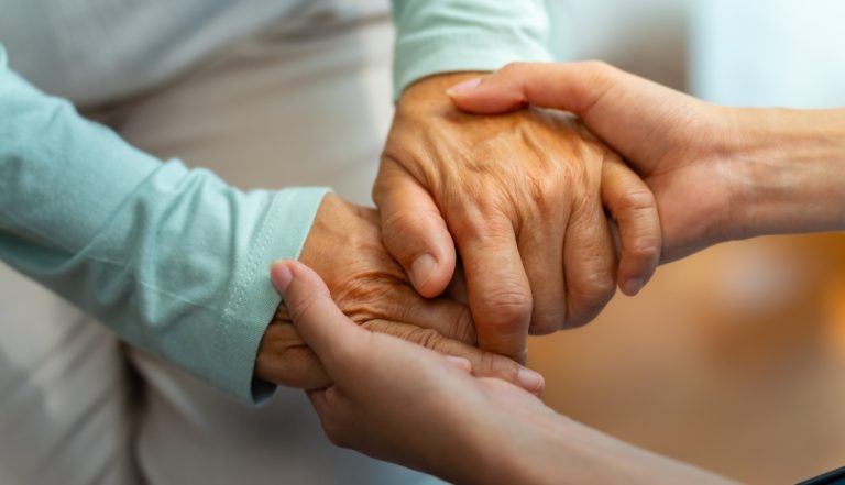 Mãos de uma mulher cuidadora a segurar mãos idosas, representando o trabalho de cuidar de idosos