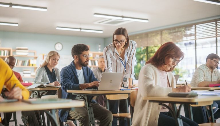 Adultos em sala de aula, acompanhados por uma professora que orienta um aluno no uso de um computador, representando o regresso às aulas após os 35 anos